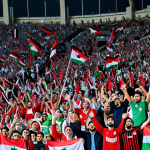 A professional image of a vibrant and diverse crowd of adult male and female fans, fully clothed in modest, national team red and white colors, enthusiastically cheering inside a large, modern football stadium. Jordanian flags wave prominently in unison as the crowd erupts in unified chants, showing immense national pride and collective spirit. The bright stadium lights illuminate the scene, capturing the electrifying atmosphere of a match day. professional photography, high quality, perfect anatomy, correct proportions, natural pose, well-formed hands, proper finger count, natural body proportions, safe for work, appropriate content, fully clothed, professional dress, family-friendly.